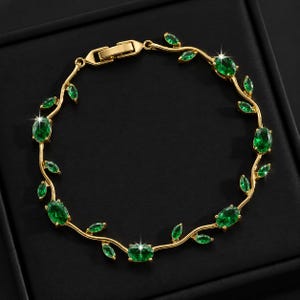 Pode incluir: Uma pulseira de tom dourado com um design de videira, apresentando pedras verdes alternadas em cortes ovais e marquise. A banda metálica imita a aparência de um ramo fino e a peça inclui um mecanismo de fecho retangular seguro.