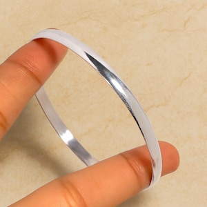 Könnte beinhalten: Ein glänzendes, silberfarbenes Armreif-Armband. Das glatte, geschwungene Design reflektiert das Licht und erzeugt ein helles, poliertes Aussehen. Das Armband wird zwischen zwei Fingern gehalten und zeigt seinen schlichten, eleganten Stil.