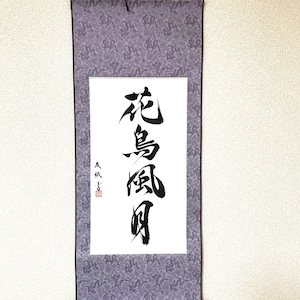 May include: A Japanese scroll with calligraphy in black ink on white paper. The scroll is framed by a patterned gray fabric and has a dark brown wooden rod at the bottom. A brown ribbon is at the top for hanging.