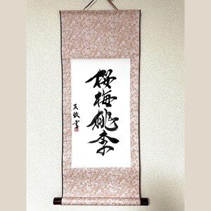 May include: A traditional Japanese scroll with calligraphy in black ink on white paper. The scroll is framed by a patterned, light pink fabric and has a dark brown wooden rod at the bottom. The top has a loop for hanging.
