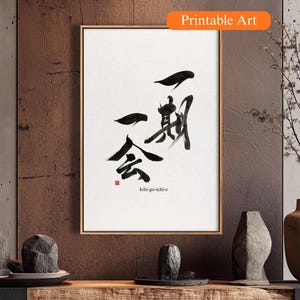 Puede incluir: Una impresión enmarcada con caligrafía japonesa en tinta negra sobre fondo blanco. La obra de arte incluye trazos de pincel y caracteres abstractos, con el texto "Ichi-go-ichi-e" en la parte inferior. El marco es dorado y la impresión está etiquetada como "Printable Art".