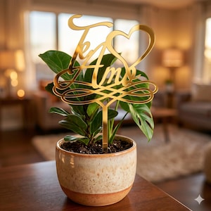 May include: A gold-colored metal plant stake with the words "Te Amo" and a heart design. The stake is inserted into a potted plant with green leaves. The pot is ceramic with a speckled pattern and a brown base.