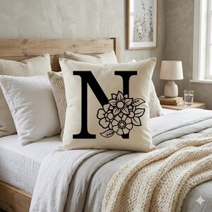May include: Cream-colored throw pillow with a large black letter "N" and floral design. The pillow is on a bed with other pillows and a beige blanket. The bed has a wooden headboard.