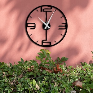 May include: A modern black wall clock with white hands and large, stylized numerals at 12, 3, 6, and 9. The clock is mounted on a pink wall, with green foliage in the foreground. The clock's hands indicate the time is approximately 10:08.
