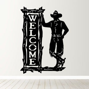 May include: Black metal sign featuring a cowboy leaning against a rustic "WELCOME" sign. The sign has a decorative wooden frame. The cowboy is wearing a hat, shirt, pants, and boots. The sign is suitable for indoor or outdoor use.