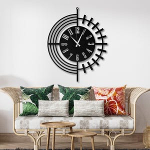 May include: Black metal wall clock with a unique design. The clock features a circular face with a minimalist style, displaying the numbers and hands. The clock is hanging on a white wall above a wicker sofa with decorative pillows.