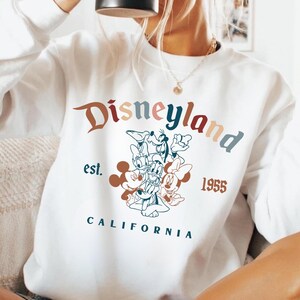 May include: White Disneyland sweatshirt with a colorful graphic featuring Mickey Mouse, Donald Duck, and Minnie Mouse. The sweatshirt has the words "Disneyland" and "California" printed on it, along with the year "1955".