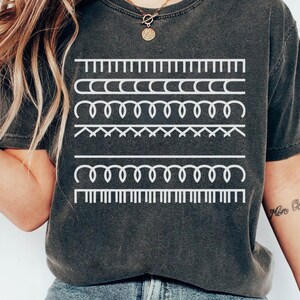 May include: Dark gray t-shirt featuring a white graphic of various sewing stitch patterns. The design includes straight lines, curved lines, and zigzags, creating a visually interesting and artistic display. The shirt is a comfortable, casual wear.