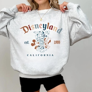 May include: Athletic gray sweatshirt with the word "Disneyland" in a retro font, along with the text "est. 1955 CALIFORNIA". The design features cartoon characters. The sweatshirt is a comfortable, casual style.