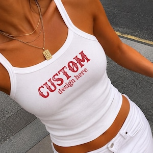 Puede incluir: Una camiseta de tirantes blanca acanalada con el texto rojo CUSTOM design here impreso en el pecho. La prenda tiene un cuello redondo y tirantes anchos. La tela presenta una textura acanalada vertical visible en toda la pieza.