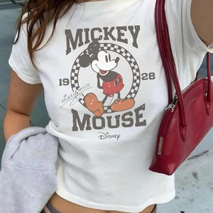 Pu&ograve; includere: T-shirt bianca con grafica vintage di Mickey Mouse, la scritta "Mickey Mouse" e "Disney". La maglia ha girocollo e maniche corte. &Egrave; visibile una borsa rossa con chiusura a zip.