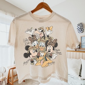 May include: Beige t-shirt featuring a graphic of Disney characters Mickey Mouse, Minnie Mouse, Donald Duck, Daisy Duck, Goofy, and Pluto. The characters are wearing safari hats. The shirt has the characters' names written in cursive.