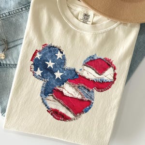 May include: Cream-coloured t-shirt with a patriotic Mickey Mouse design. The design incorporates denim and red fabric, with white stars on a blue background, resembling the American flag. The shirt is paired with blue jeans and a brown hat.
