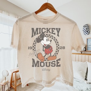 May include: Cream-colored t-shirt featuring a distressed graphic of Mickey Mouse. The design includes the words "Mickey Mouse" and "Disney" along with the years "1928" and "19". The shirt is hanging on a wooden hanger.