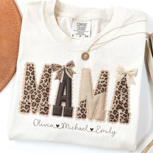 May include: Cream-colored Comfort Colors t-shirt with the word "MAMA" in leopard print and brown letters. The shirt has bows and the names "Olivia", "Michael", and "Emily" below. A gold necklace and a brown hat are also visible.