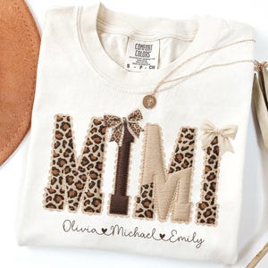May include: Cream-coloured t-shirt featuring the word "MIMI" in leopard print and brown fabric letters, decorated with bows. The names "Olivia", "Michael", and "Emily" are printed below. A gold necklace and a brown hat are also visible.
