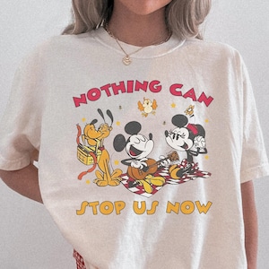 May include: Cream-colored t-shirt featuring a graphic of Mickey Mouse, Minnie Mouse, and Pluto, with the text "NOTHING CAN STOP US NOW" in red and yellow. The characters are depicted on a picnic blanket. The person is wearing red and white checkered shorts.