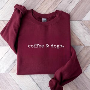 May include: Maroon crewneck sweatshirt with the text "coffee & dogs." The sweatshirt is made of a soft material and is displayed on a light wood grain surface. A maroon circle with the word "maroon" is in the upper right corner.