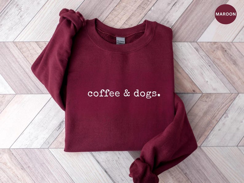 May include: Maroon crewneck sweatshirt with the text "coffee & dogs." The sweatshirt is made of a soft material and is displayed on a light wood grain surface. A maroon circle with the word "maroon" is in the upper right corner.