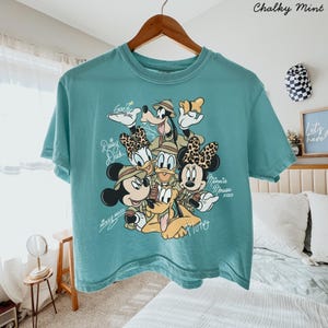 May include: A teal-colored t-shirt featuring a graphic of Disney characters, including Mickey Mouse, Minnie Mouse, Donald Duck, Daisy Duck, Goofy, and Pluto. The text "Mickey Mouse" and "Minnie Mouse xoxo" are also visible. The shirt is hanging on a wooden hanger.