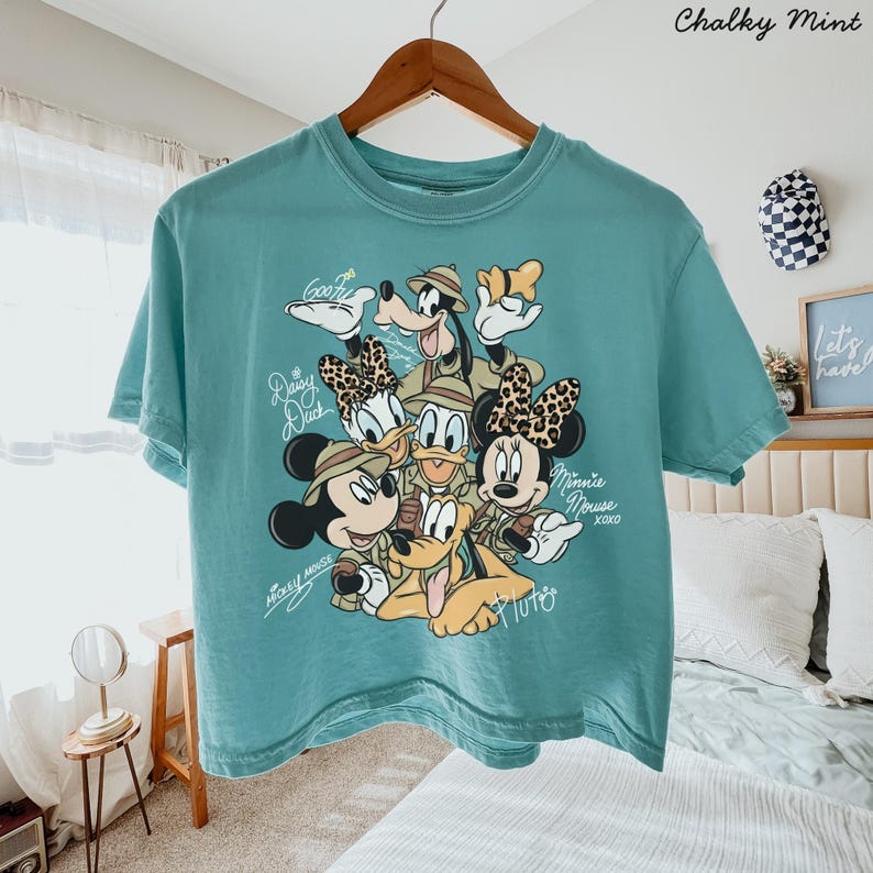 May include: A teal-colored t-shirt featuring a graphic of Disney characters, including Mickey Mouse, Minnie Mouse, Donald Duck, Daisy Duck, Goofy, and Pluto. The text "Mickey Mouse" and "Minnie Mouse xoxo" are also visible. The shirt is hanging on a wooden hanger.