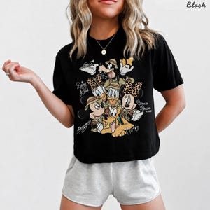 May include: Black t-shirt featuring a graphic of Disney characters Mickey Mouse, Minnie Mouse, Donald Duck, Daisy Duck, Goofy, and Pluto. The characters are wearing safari hats and animal print accents. Text includes "Mickey Mouse", "Daisy Duck", "Goofy", "Minnie Mouse XOXO", and "Pluto".