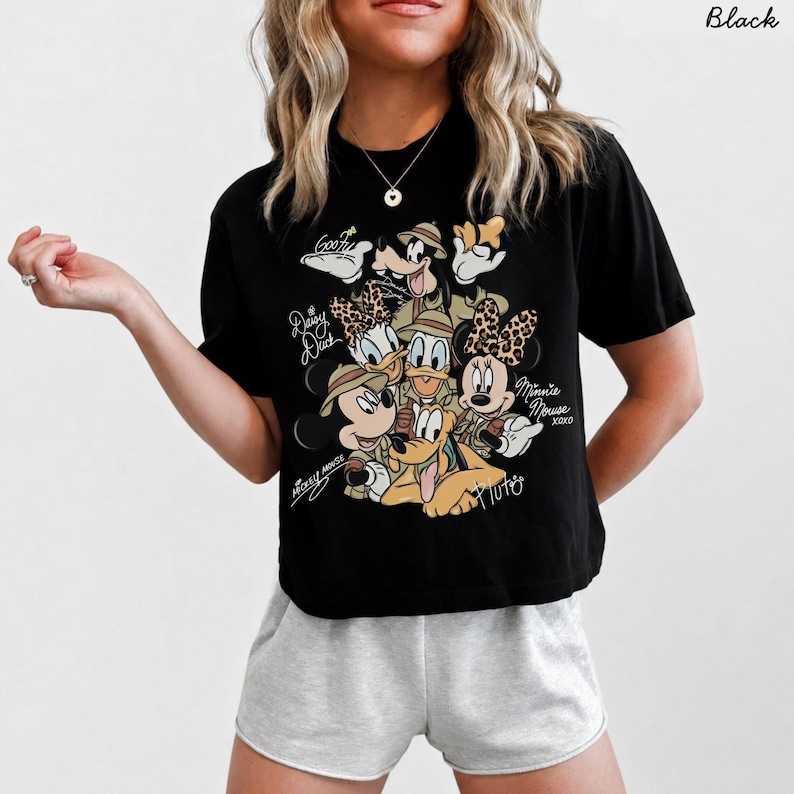 May include: Black t-shirt featuring a graphic of Disney characters Mickey Mouse, Minnie Mouse, Donald Duck, Daisy Duck, Goofy, and Pluto. The characters are wearing safari hats and animal print accents. Text includes "Mickey Mouse", "Daisy Duck", "Goofy", "Minnie Mouse XOXO", and "Pluto".