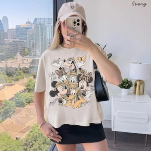 May include: Ivory-colored t-shirt featuring a graphic of Disney characters Mickey Mouse, Minnie Mouse, Donald Duck, Daisy Duck, Goofy, and Pluto. The characters are wearing safari outfits. The shirt is paired with a black skirt and a beige cap.