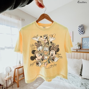 May include: Yellow short-sleeved t-shirt featuring a graphic of Disney characters Mickey Mouse, Minnie Mouse, Donald Duck, Daisy Duck, Goofy, and Pluto. The characters are wearing safari hats. The text "Mickey Mouse" and "Minnie Mouse" are written on the shirt.