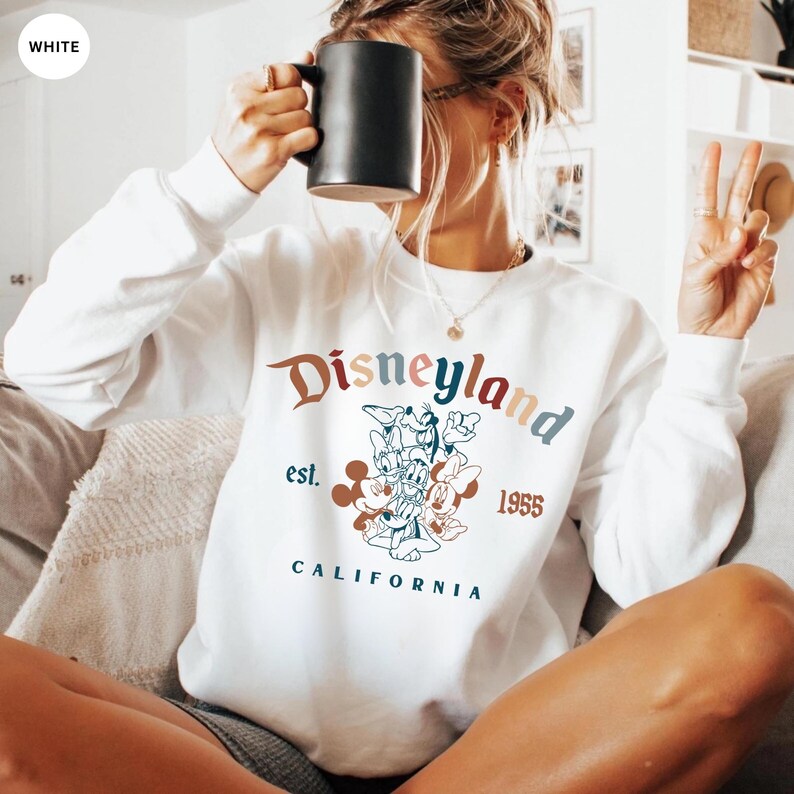 May include: White Disneyland sweatshirt with a colorful graphic featuring Mickey Mouse, Donald Duck, and Minnie Mouse. The sweatshirt has the words "Disneyland" and "California" printed on it, along with the year "1955".