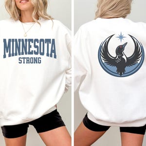 May include: White sweatshirt with the text "MINNESOTA STRONG" in blue block letters. The back of the sweatshirt features a graphic of a bird with outstretched wings inside a blue circle, with a star above.
