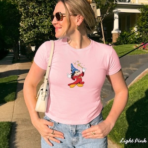 Może przedstawiać: Jasnor&oacute;żowa koszulka z kresk&oacute;wkowym wizerunkiem Myszki Miki przebranej za czarodzieja, trzymającej magiczną r&oacute;żdżkę. Koszulka jest połączona z niebieskimi dżinsami. Słowa "Light Pink" są wydrukowane w prawym dolnym rogu.