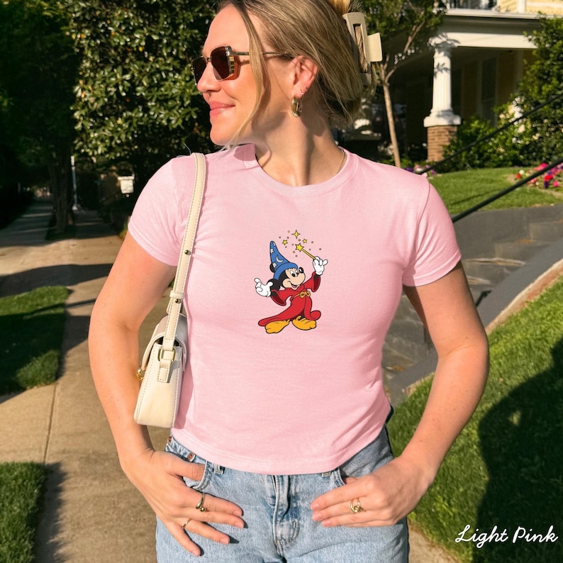 Może przedstawiać: Jasnor&oacute;żowa koszulka z kresk&oacute;wkowym wizerunkiem Myszki Miki przebranej za czarodzieja, trzymającej magiczną r&oacute;żdżkę. Koszulka jest połączona z niebieskimi dżinsami. Słowa "Light Pink" są wydrukowane w prawym dolnym rogu.