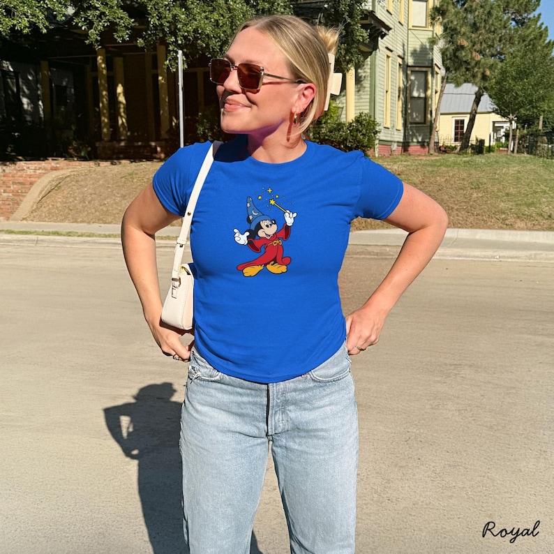 Może przedstawiać: Kr&oacute;lewski niebieski t-shirt z grafiką Myszki Miki jako Ucznia Czarodzieja. Postać z kresk&oacute;wki jest w czerwonej szacie i niebieskim kapeluszu, trzymając r&oacute;żdżkę z ż&oacute;łtą gwiazdą. Koszulka jest sparowana z jasnoniebieskimi dżinsami.