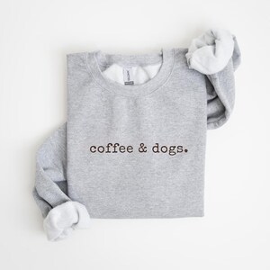 May include: Athletic gray sweatshirt with the text "coffee & dogs." The sweatshirt has a crew neck and long sleeves. The sleeves are cuffed, and the inside of the cuffs are white. A circular tag in the upper right corner reads "ATHLETIC GRAY."