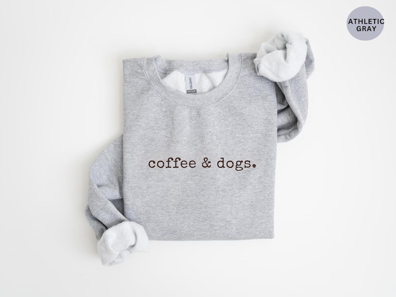 May include: Athletic gray sweatshirt with the text "coffee & dogs." The sweatshirt has a crew neck and long sleeves. The sleeves are cuffed, and the inside of the cuffs are white. A circular tag in the upper right corner reads "ATHLETIC GRAY."