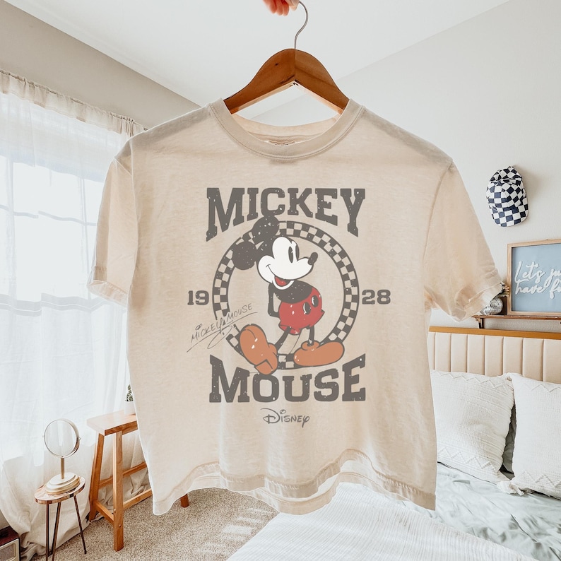 May include: Cream-colored t-shirt featuring a distressed graphic of Mickey Mouse. The design includes the words "Mickey Mouse" and "Disney" along with the years "1928" and "19". The shirt is hanging on a wooden hanger.