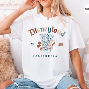 May include: White t-shirt with a Disneyland graphic. The design features the word "Disneyland" in a multi-colored font, the year "1955", and the word "California". A floral illustration of Disney characters is also included.