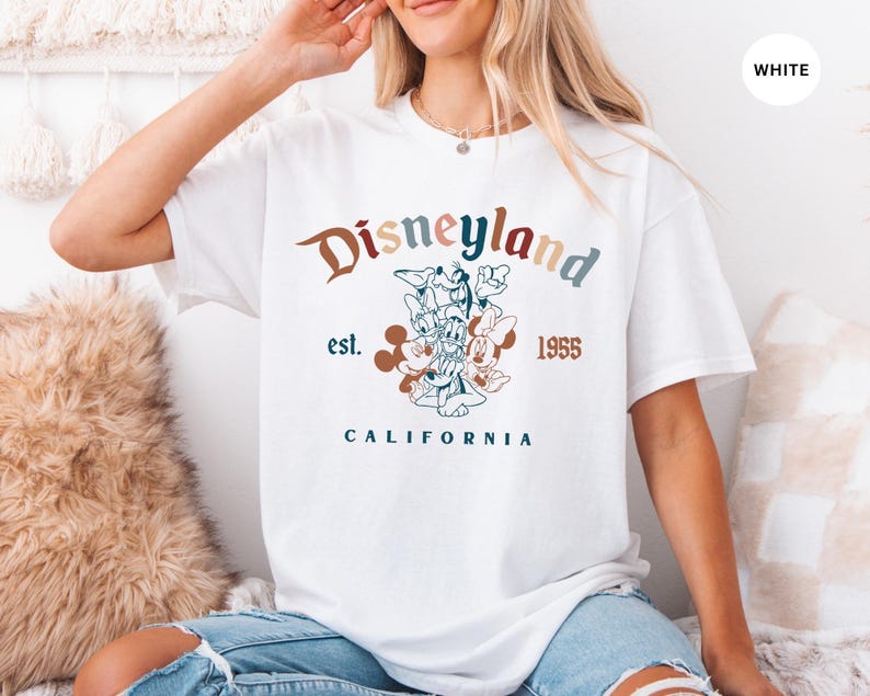 May include: White t-shirt with a Disneyland graphic. The design features the word "Disneyland" in a multi-colored font, the year "1955", and the word "California". A floral illustration of Disney characters is also included.