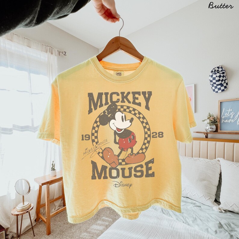 May include: Yellow short-sleeve t-shirt featuring a vintage Mickey Mouse graphic. The design includes the words "Mickey Mouse" and the year "1928" in a circular checkered pattern. The shirt is held up by a wooden hanger.