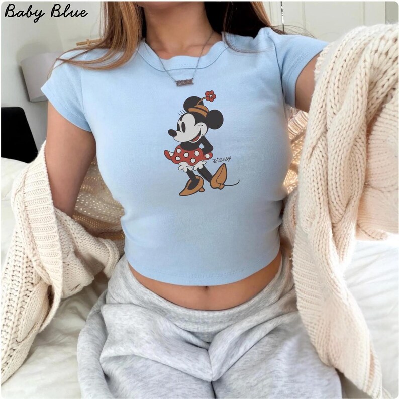 May include: Pale blue crop top with a Minnie Mouse design. The graphic depicts Minnie Mouse in a red polka dot skirt, black shoes, and a flower on her head. The top has short sleeves and the text "Baby Blue" is in the upper left corner.