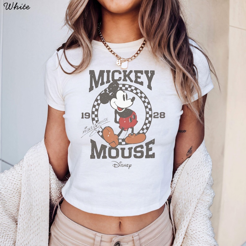 Pu&ograve; includere: Top corto bianco con grafica vintage di Mickey Mouse. Il design include le parole "Mickey Mouse" e l'anno "1928" in una cornice circolare a scacchi. Il logo Disney &egrave; in basso.