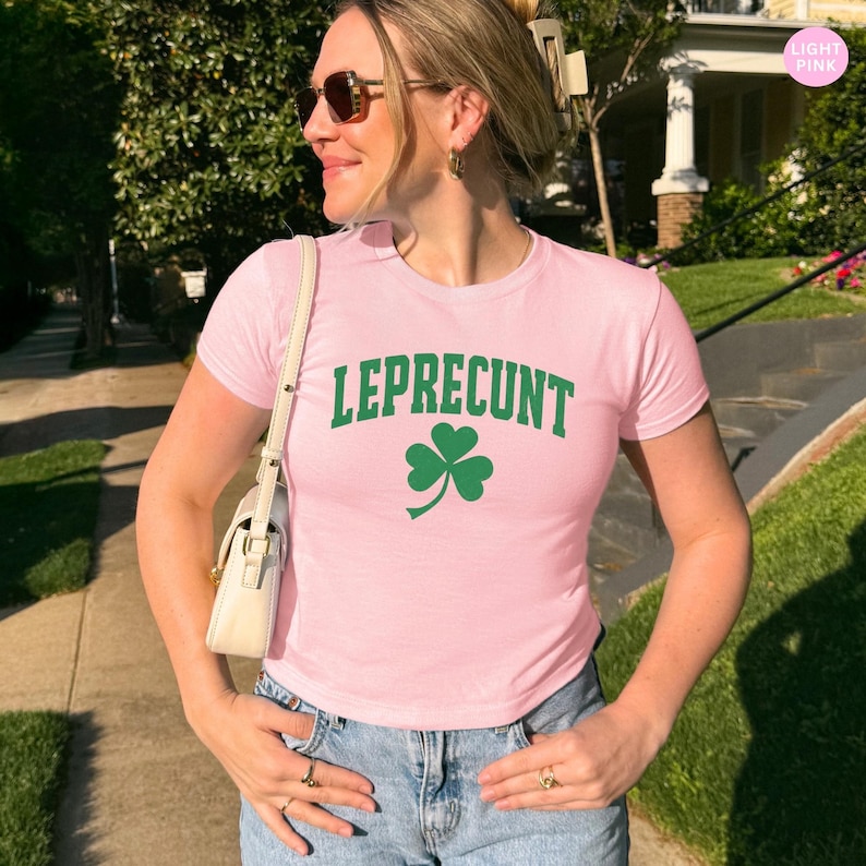 May include: Light pink t-shirt with the word "LEPRECUNT" in green block letters and a green shamrock graphic. The shirt is short-sleeved and made of a soft material. The model is wearing sunglasses and blue jeans.