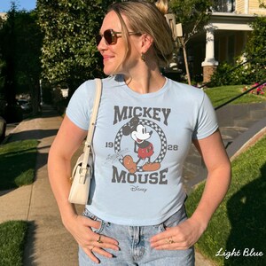 Pu&ograve; includere: T-shirt azzurra con grafica vintage di Topolino. Il design include il testo "Mickey Mouse" e gli anni "1928" e "1928". La maglietta &egrave; abbinata a jeans blu e una borsa color crema.
