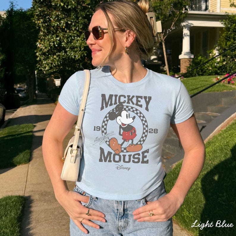 Pu&ograve; includere: T-shirt azzurra con grafica vintage di Topolino. Il design include il testo "Mickey Mouse" e gli anni "1928" e "1928". La maglietta &egrave; abbinata a jeans blu e una borsa color crema.