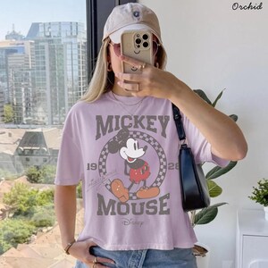 May include: A light purple t-shirt featuring a graphic of Mickey Mouse. The design includes the words "Mickey Mouse" and the years "19" and "28". The shirt is a cropped style and has short sleeves. A beige baseball cap and black purse are also visible.