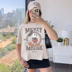 May include: Cream-colored t-shirt featuring a distressed Mickey Mouse graphic. The design includes the words "Mickey Mouse" and the years "1928" and "1928". The shirt is paired with a black skirt and a beige baseball cap.