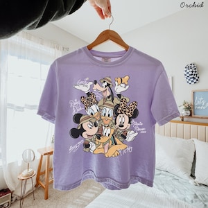 May include: A lavender t-shirt featuring a graphic of Disney characters Mickey Mouse, Minnie Mouse, Donald Duck, Daisy Duck, Goofy, and Pluto. The characters are wearing safari outfits. The word "Orchid" is in the top right corner.