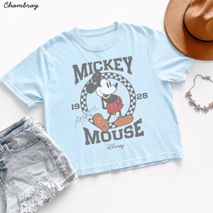 May include: Light blue t-shirt featuring a distressed graphic of Mickey Mouse with the text "Mickey Mouse 1928". The shirt is paired with denim shorts, a brown hat, and a floral necklace. Disney apparel.