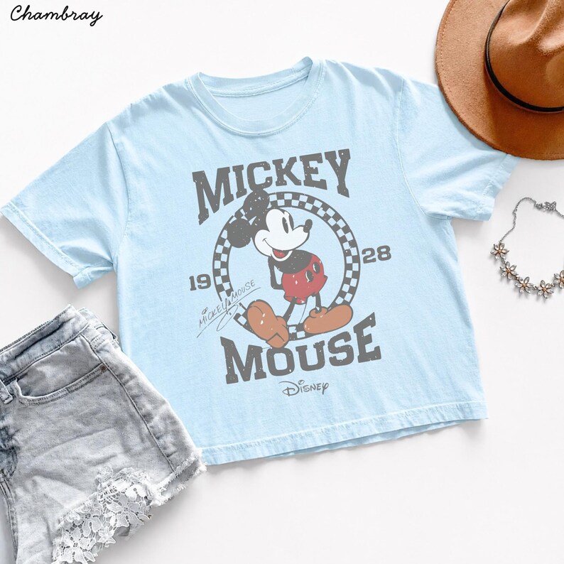May include: Light blue t-shirt featuring a distressed graphic of Mickey Mouse with the text "Mickey Mouse 1928". The shirt is paired with denim shorts, a brown hat, and a floral necklace. Disney apparel.